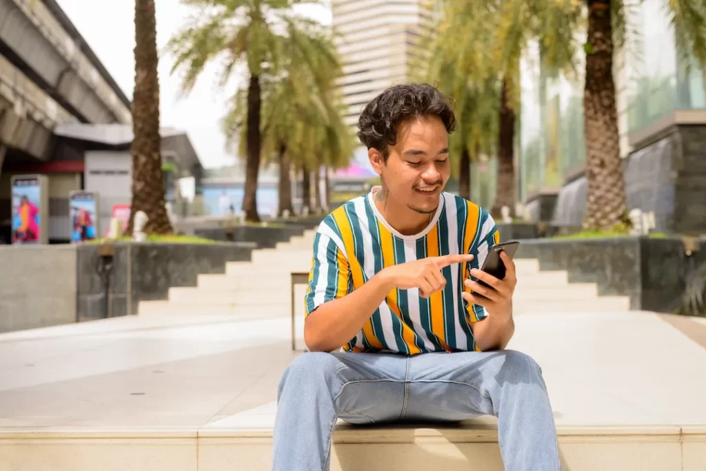 Portrait of happy multi-ethnic young man wearing colorful shirt outdoors during summer while using mobile phone